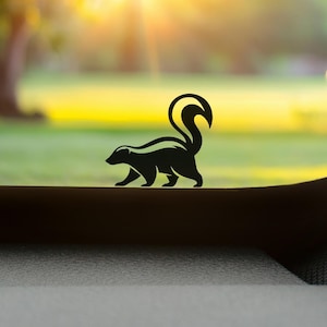 May include: A black vinyl decal featuring a stylised silhouette of a skunk with its tail arched upwards. The design shows the animal in a walking profile, characterised by its distinct striped pattern and curved tail, applied to a smooth surface.