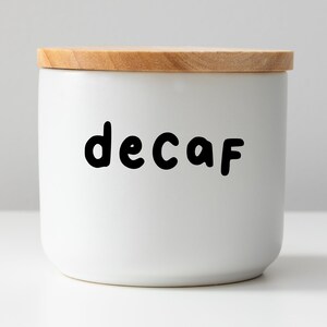 Puede incluir: Un tarro de cerámica blanco con tapa de madera, etiquetado con "decaf" en letras negras.