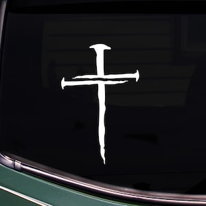 May include: White, stylized cross sticker with a nail-like design. The cross is set against a dark background, possibly a car window. The design has a textured, hand-drawn appearance, suggesting a religious theme.