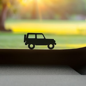 May include: A black silhouette of a small SUV with round wheels and a spare tyre on the back. The vehicle is set against a blurred background of green grass, trees, and a bright yellow sky.