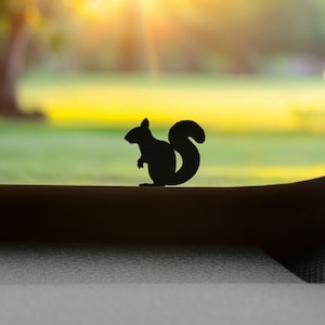 Könnte beinhalten: Schwarze Silhouette eines Eichhörnchens mit buschigem Schwanz. Das Eichhörnchen sitzt auf einer dunklen Oberfläche, vor einem verschwommenen grünen und gelben Hintergrund eines Parks. Sonnenschein.