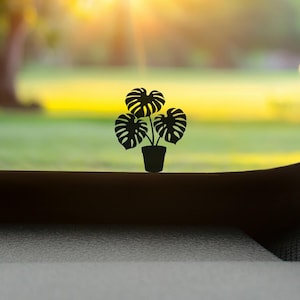 May include: Silhouette of a potted Monstera plant with large, split leaves. The black plant is set against a blurred background of green grass and trees, with bright sunlight shining through. The plant is in a black pot.