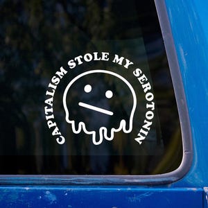 May include: White vinyl sticker with the text "CAPITALISM STOLE MY SEROTONIN" in a circular design around a sad-faced, melting blob graphic. The sticker is affixed to a blue vehicle window.