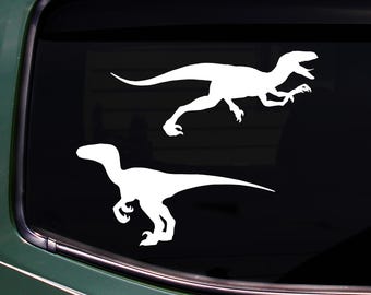 May include: Two white dinosaur decals on a black background. The top decal shows a running raptor with its mouth open. The bottom decal is a raptor in a similar pose. The decals are likely for a car window.