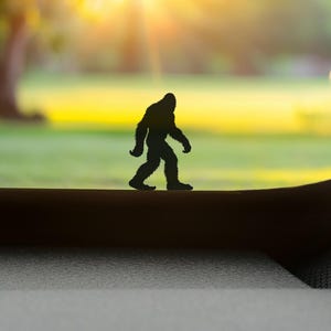 May include: A black silhouette of a Bigfoot figure walking across a dark surface, with a blurred background of green grass and a bright yellow sky. The figure is a dark colour and appears to be made of plastic or a similar material.