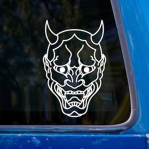Peut inclure: Autocollant en vinyle blanc représentant un masque Oni japonais. Le design présente le visage du démon avec des cornes, des yeux et une large bouche. L'autocollant est affiché sur une surface bleue.