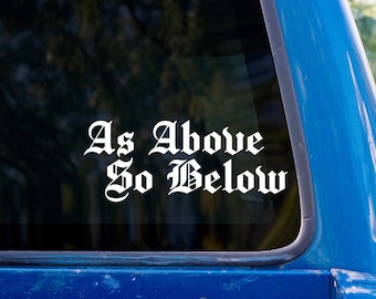May include: White vinyl decal with the phrase "As Above So Below" in a gothic font, applied to a blue vehicle window. The background shows a blurred view of trees.