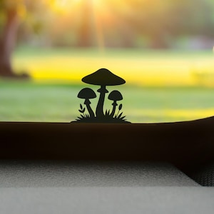 May include: A silhouette of three mushrooms with grass and leaves is set against a blurred outdoor background. The mushrooms are in varying sizes, with a large one in the center and two smaller ones on either side. The background features a green lawn and a bright, sunny sky.