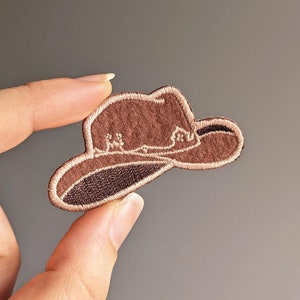 May include: A brown felt cowboy hat with a light brown embroidered design. The hat is a patch or applique that can be ironed onto fabric.