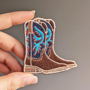 Puede incluir: Un par de botas vaqueras marrones con bordados azules y naranjas. Las botas están cosidas en un parche de tela.