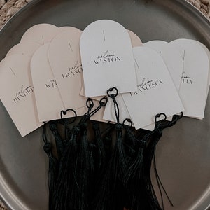 May include: A set of ten cream-coloured arched cards with black text that says "welcome" followed by a name. The cards are tied together with black tassels.