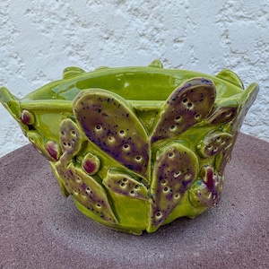 May include: A lime green ceramic planter featuring a three-dimensional relief of prickly pear cactus pads. The cactus pads are accented with purple and dark blue speckles and small indentations, creating a textured surface around the bowl.