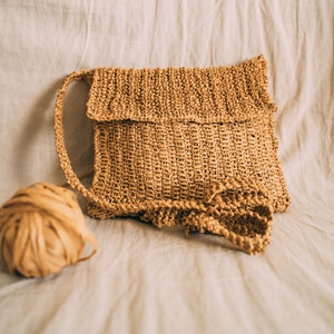 May include: A light brown woven crossbody bag with a flap closure. The bag is made of a natural material and has a casual, bohemian style.