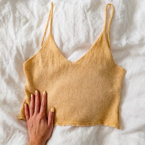 Könnte beinhalten: Ein hellgelbes Stricktop mit dünnen Trägern. Das Top liegt flach auf einer weißen Leinenoberfläche.