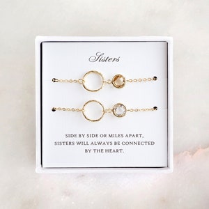 Könnte beinhalten: Zwei goldene Armbänder mit einem Kreis-Anhänger und einem kleinen Edelstein. Die Armbänder befinden sich in einer weißen Schachtel mit dem Text "Sisters" und "Seite an Seite oder meilenweit entfernt, Schwestern werden immer durch das Herz verbunden sein."