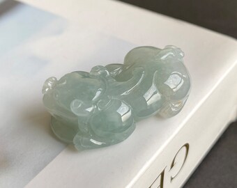 May include: A jade pendant carved in the shape of two mythical creatures. The pendant is a light green color and has a smooth, polished finish.