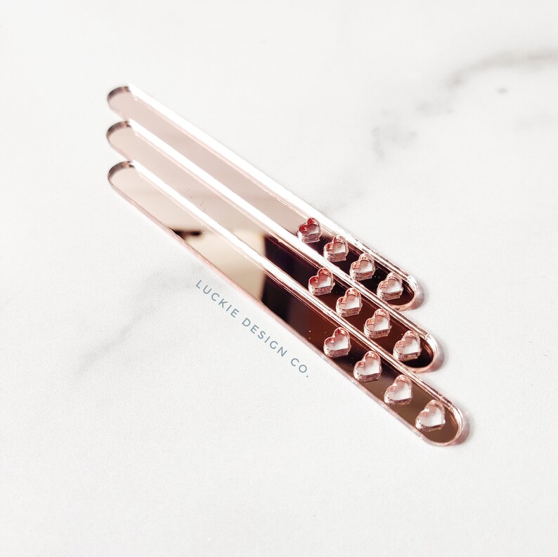 May include: Three rose gold acrylic sticks with heart cutouts. The sticks are stacked on top of each other. The text "LUCKIE DESIGN CO." is printed on the bottom stick.