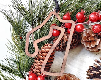 May include: A wooden Christmas ornament shaped like a race track, with a brown finish and a black string for hanging. The ornament is surrounded by pine branches and red berries.