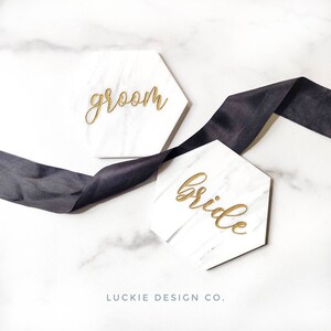 May include: Two white marble hexagonal coasters with gold lettering. The top coaster says "groom" and the bottom coaster says "bride".