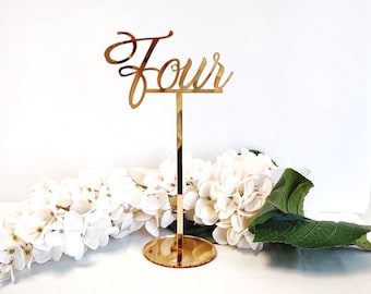 May include: Gold mirror acrylic table number with the word "Four" in a cursive font. The number is attached to a gold metal stand.