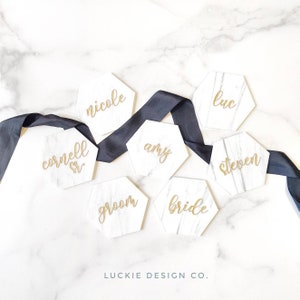 May include: White hexagonal marble place cards with gold calligraphy names, including "bride", "groom", "amy", "luc", "nicole", "cornell sr", and "steven". The place cards are arranged on a white marble surface with a black ribbon.