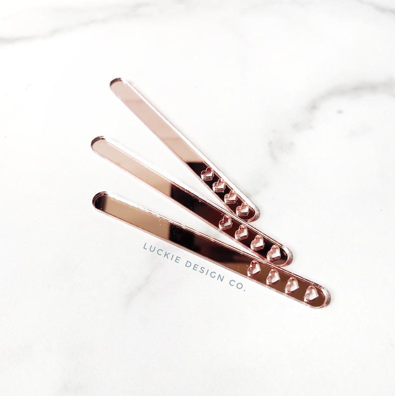 May include: Three rose gold acrylic bookmark strips with a row of small heart cutouts. The bookmarks are stacked on top of each other and the text "LUCKIE DESIGN CO." is printed on the white surface below.