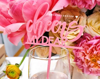 May include: Pink acrylic cheers my dear sign with a pink stem, perfect for a celebratory drink. The sign is in front of a bouquet of pink and white flowers.