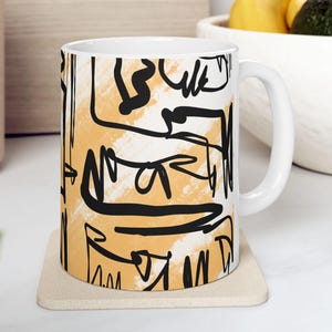 May include: A white ceramic mug featuring a bold abstract design with thick black hand-drawn lines layered over a soft, textured mustard yellow brushstroke background. The mug has a standard cylindrical shape with a curved handle on the right side.