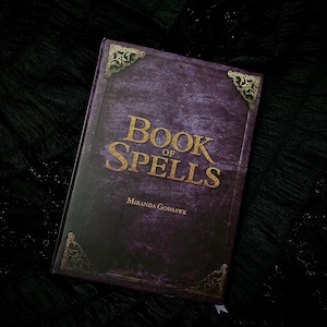 Może przedstawiać: Fioletowa książka z złotymi akcentami i tytułem "Book of Spells" autorstwa Mirandy Goshawk.