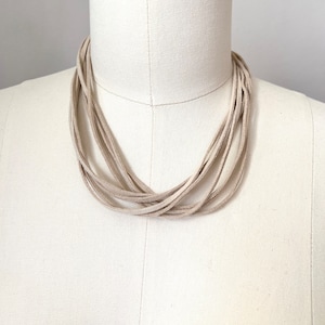 May include: A multi-strand necklace made of light beige suede leather. The necklace is layered and has a simple, minimalist design.