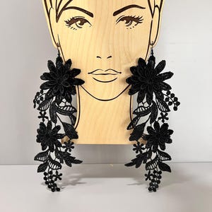 Puede incluir: Un par de pendientes largos de encaje negro con diseños florales. Los pendientes son colgantes y presentan delicadas flores y hojas de encaje negro.
