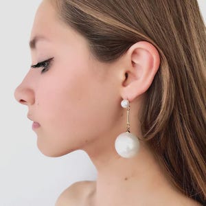 May include: A pair of elegant earrings featuring a small pearl stud and a long, thin gold bar suspending a large, round white pearl. The earrings are worn by a person with long brown hair.