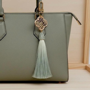 Peut inclure: Un sac à main vert sauge avec une fermeture éclair et des accessoires dorés. Un charm décoratif doré en forme de trèfle avec un pompon est suspendu à la poignée. Le pompon est de couleur vert clair.