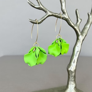 Op de afbeelding: Een paar limoengroene bloemvormige oorbellen met goudkleurige haken. De oorbellen worden gepresenteerd op een zilverkleurige decoratieve tak. De oorbellen hebben meerdere bloemblaadjes en een heldere kleur.