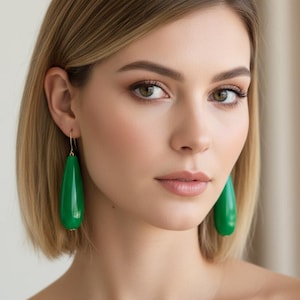 May include: A pair of drop earrings featuring smooth, glossy, teardrop-shaped green pendants suspended from thin gold-toned metal hooks. The earrings have a polished finish and a simple, elongated design that hangs vertically from the earlobes.