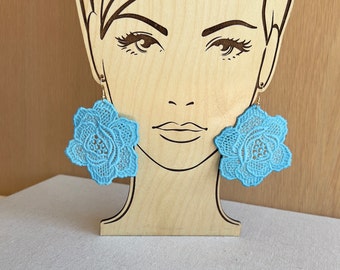 May include: A pair of blue lace flower earrings with gold hooks. The earrings are displayed on a wooden mannequin head.