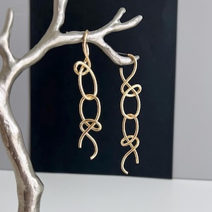 May include: A pair of gold-colored dangle earrings. Each earring features a series of connected oval links and a looped design. The earrings are displayed against a black and silver background.