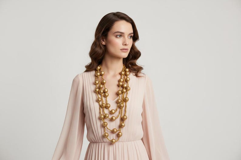 May include: A gold-colored beaded necklace with multiple strands of large, round beads. The necklace is worn over a light pink dress with long sleeves. The background is a plain, light gray.