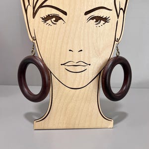 May include: A pair of dark brown wooden hoop earrings with a brass hook. The earrings are displayed on a wooden mannequin head.