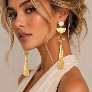 May include: Gold-tone drop earrings featuring a hexagonal white stone stud, a semi-circular textured piece, a series of concentric circles, and a long, hammered teardrop-shaped pendant. The earrings have a metallic finish and a multi-layered design.