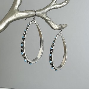May include: A pair of silver-toned teardrop earrings with a distressed finish. Each earring is adorned with small, light blue beads along the outer edge. The earrings are suspended from a silver-colored decorative branch.
