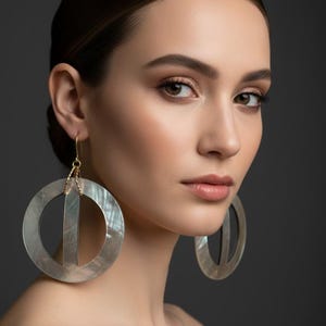 May include: Large circular earrings featuring a flat, iridescent mother-of-pearl disc with a central vertical cutout. The earrings are suspended from thin, gold-toned metal hooks that attach to the top of the circular frame with a small link.