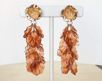 May include: A pair of long, dangling earrings with a gold flower charm at the top and a cascade of brown, translucent petals hanging below.