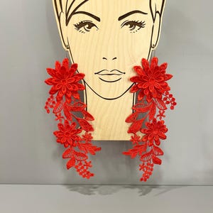 Puede incluir: Un par de pendientes largos de encaje floral rojo. Los pendientes son colgantes y presentan una flor grande en la parte superior y flores y hojas más pequeñas que caen en cascada.