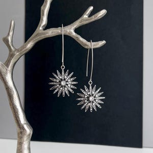 May include: A pair of silver-toned starburst earrings with a central clear stone and surrounding smaller stones. The earrings hang from long, thin wires. The earrings are displayed on a silver decorative branch against a black and white background.