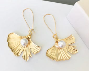 May include: A pair of gold-toned earrings featuring a ginkgo leaf-shaped charm with a single white pearl accent. The earrings are suspended from long, thin gold-toned wire hooks that loop through the top of each metallic leaf-shaped pendant.