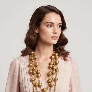 May include: A woman wearing a multi-strand gold beaded necklace. The necklace features large, round beads and hangs in multiple layers. The woman is wearing a light pink top. The background is a solid, neutral color.