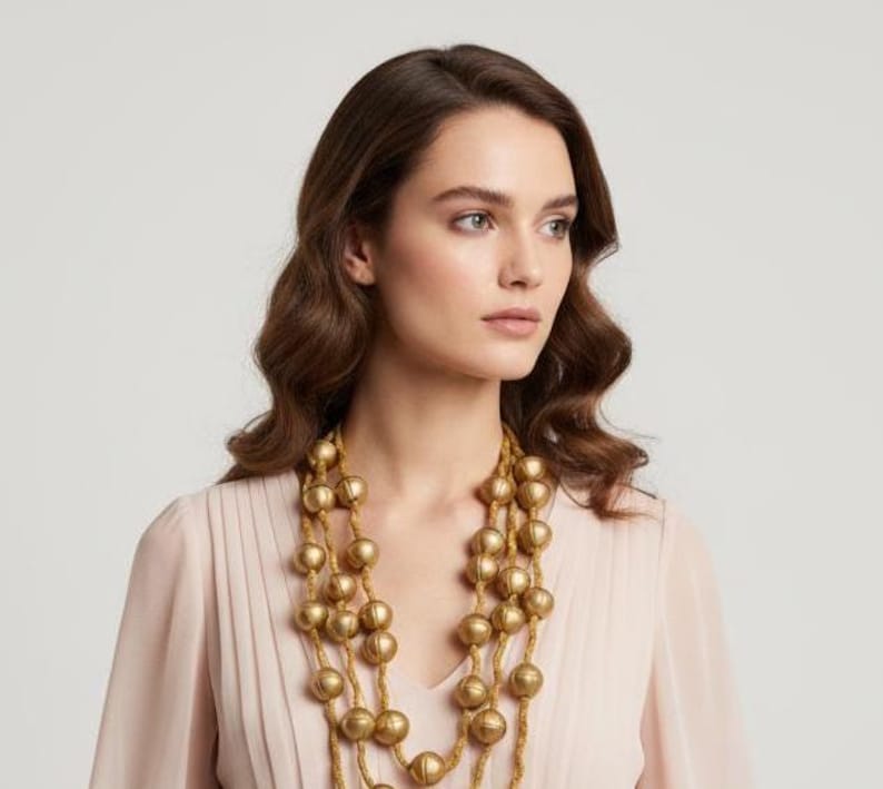 May include: A woman wearing a multi-strand gold beaded necklace. The necklace features large, round beads and hangs in multiple layers. The woman is wearing a light pink top. The background is a solid, neutral color.