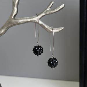 May include: A pair of drop earrings featuring spherical black beads with a faceted, textured surface. The beads are suspended from long, slender silver-toned metal hooks that curve into a simple, elongated arch shape, creating a dangling effect.