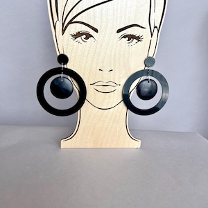 May include: Black, oversized hoop earrings with a circular design. The earrings are displayed on a wooden stand shaped like a woman's face. The earrings have a large outer circle and a smaller inner circle, with a small circular accent at the top.